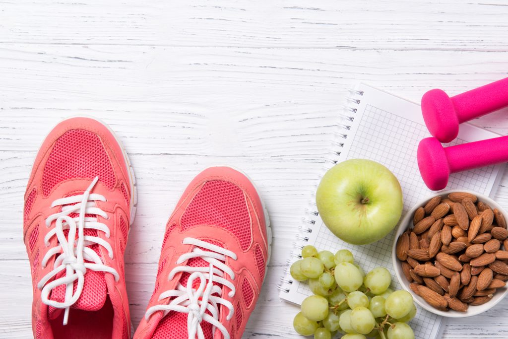 Fitness and healthy eating concept, pink sneakers and pink dumbbells with apple, grapes and almond nuts on notepad, wooden background, top view with copy space